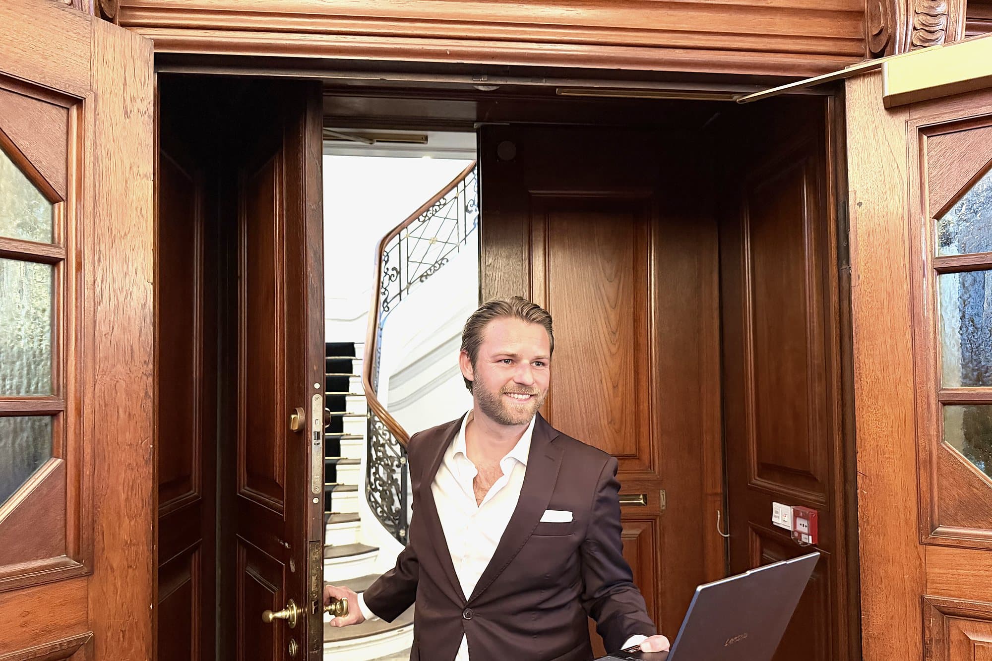 Kim Olsen smiling in an open wooden doorway with a laptop in one hand and a spiral staircase behind him.