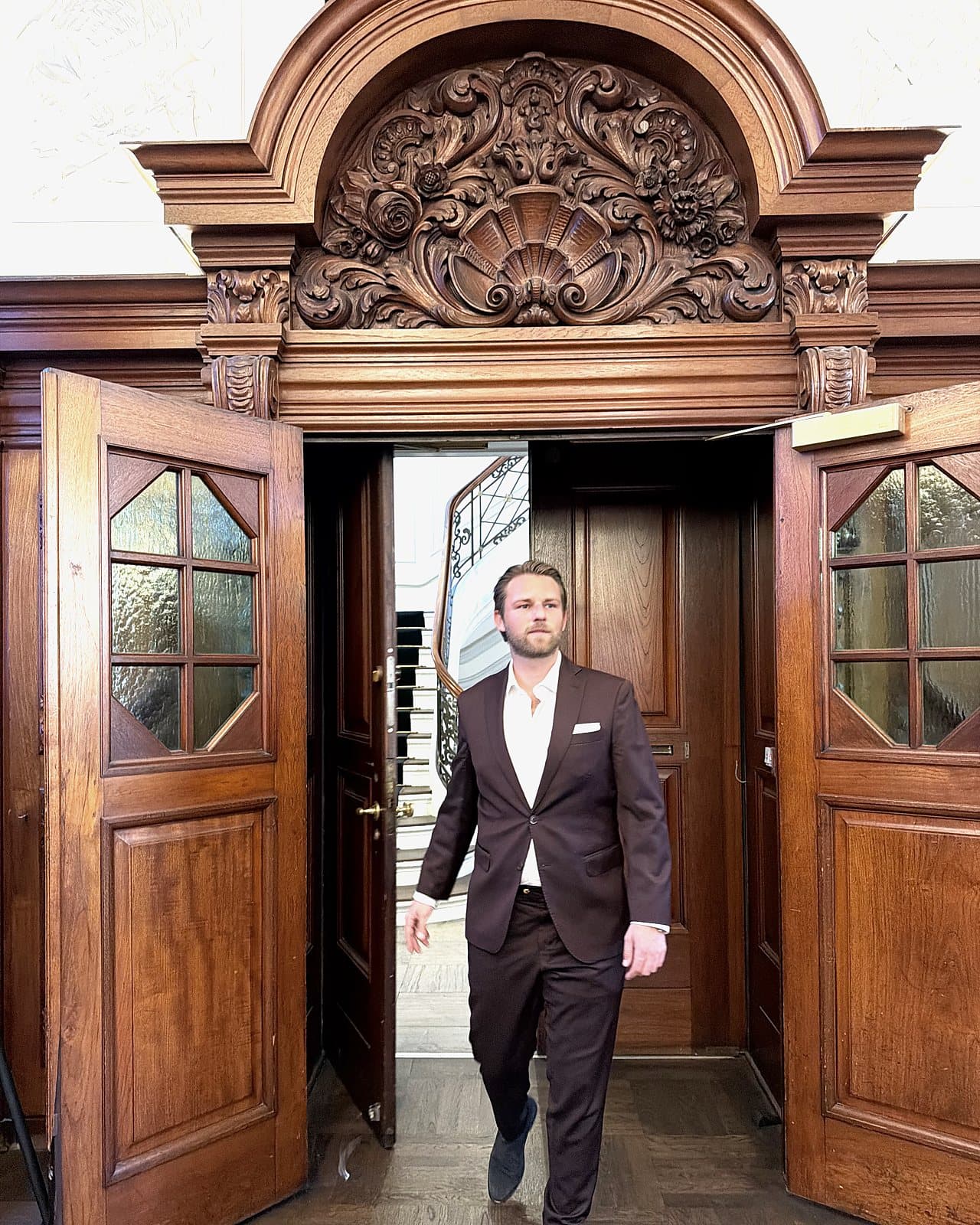 Kim Olsen in a dark suit walking through a pair of open, carved wooden double doors, with a spiral staircase behind him.