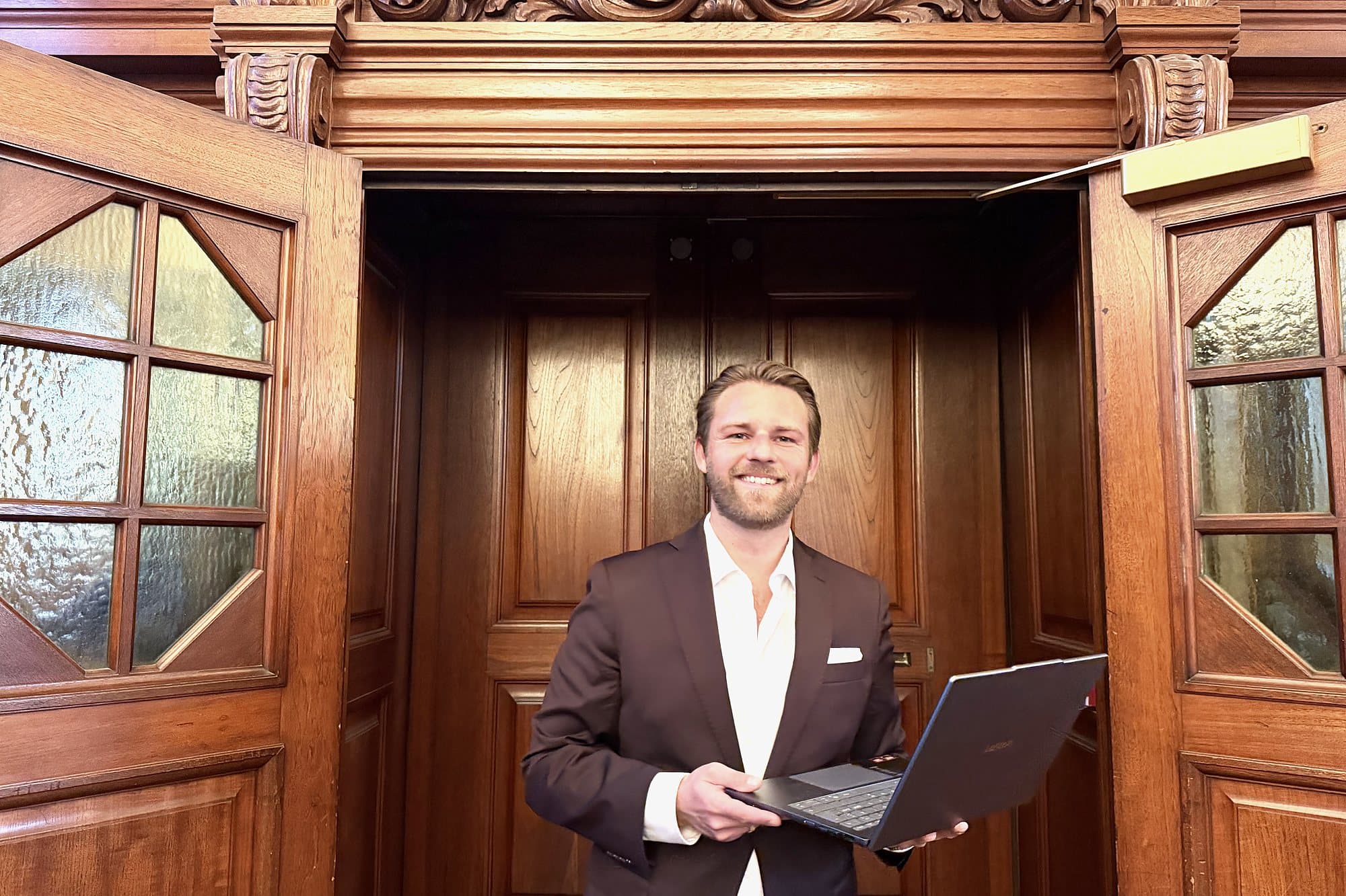 Kim Olsen smiling in a pair of open, carved wooden doors, holding a laptop.