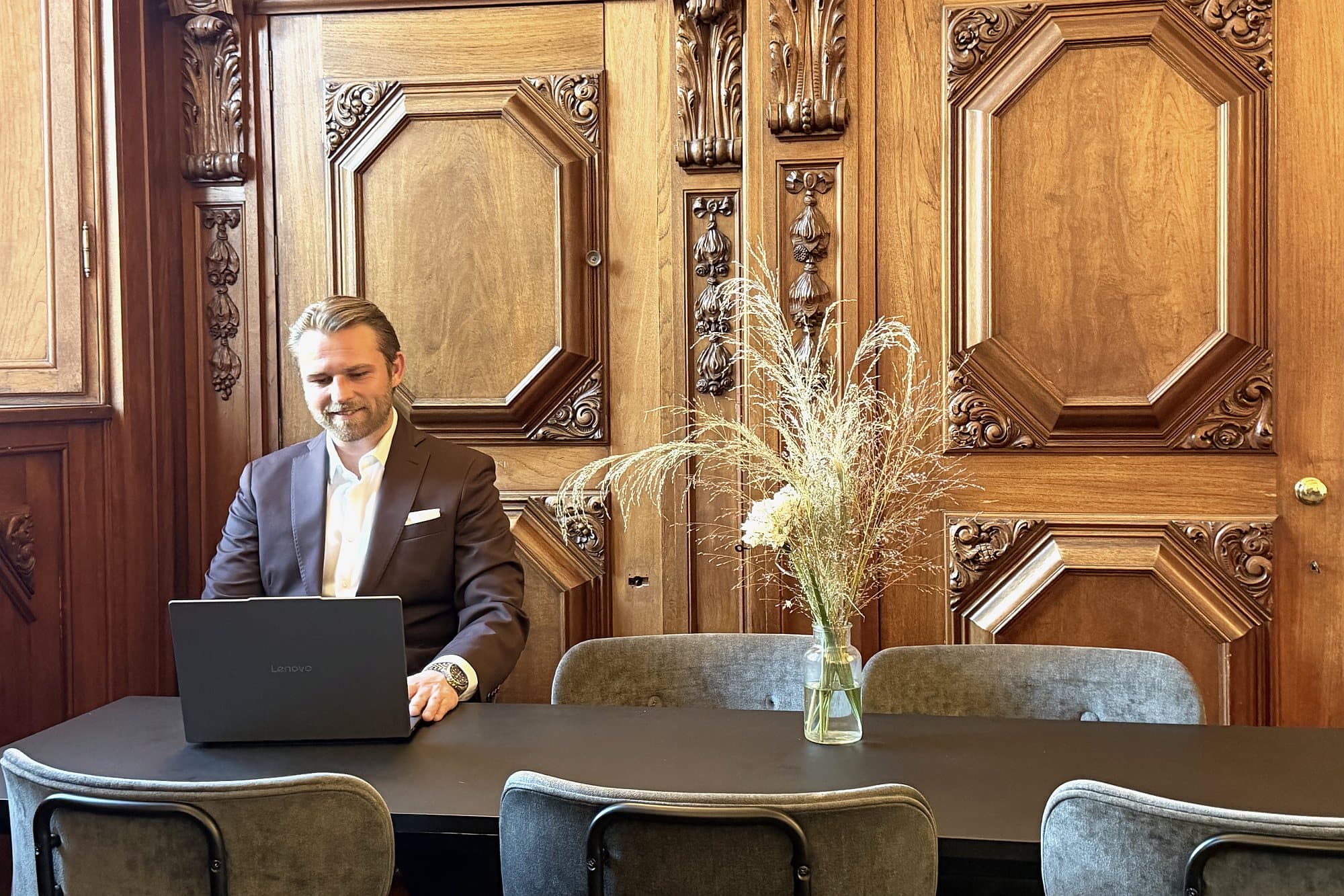 Kim Olsen working on a laptop at a long black table, with dried flowers in a vase and carved panelling behind.