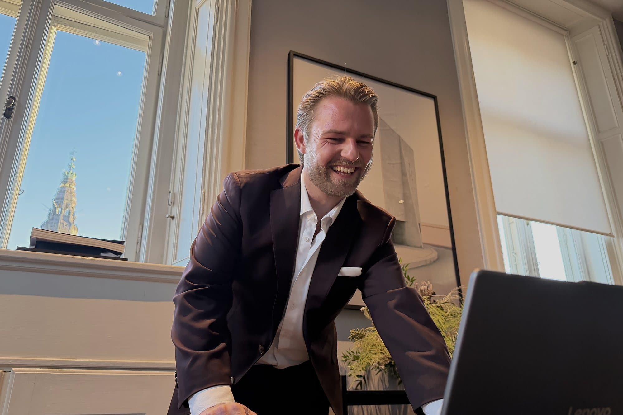 Kim Olsen laughing as he leans over an open laptop by a tall window with an old copper spire outside.