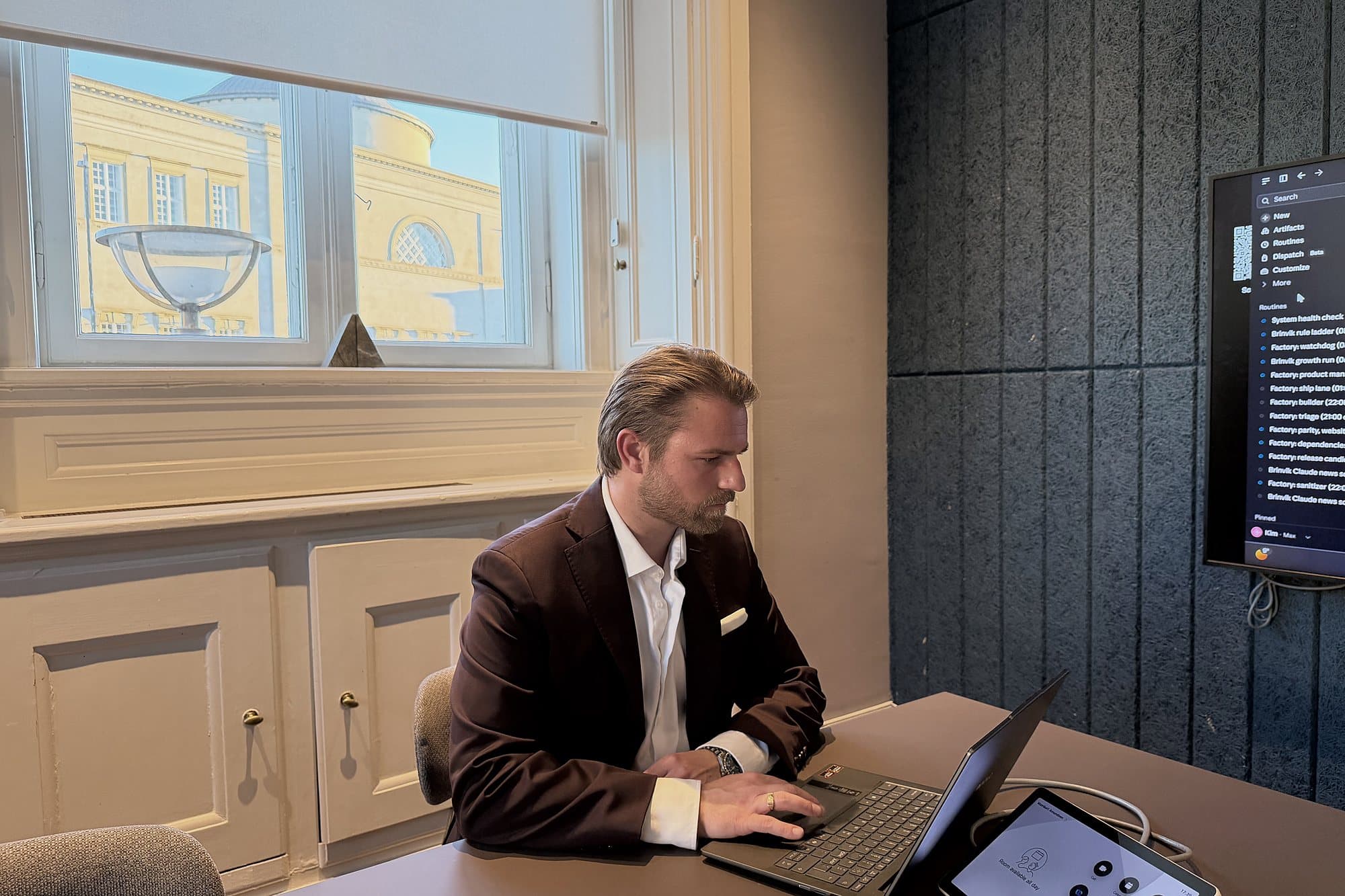 Kim Olsen working on a laptop at a meeting table, with a list of Claude routines on the screen on the wall beside him.