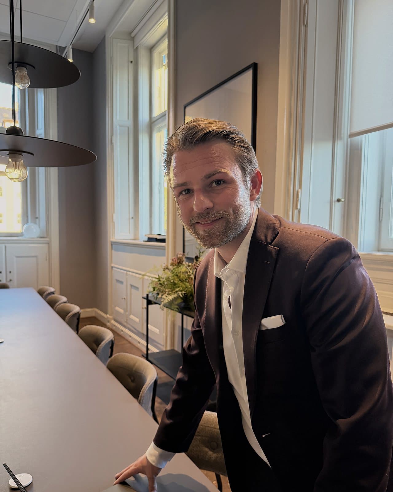 Kim Olsen in a dark suit, leaning on a long meeting table and smiling at the camera.