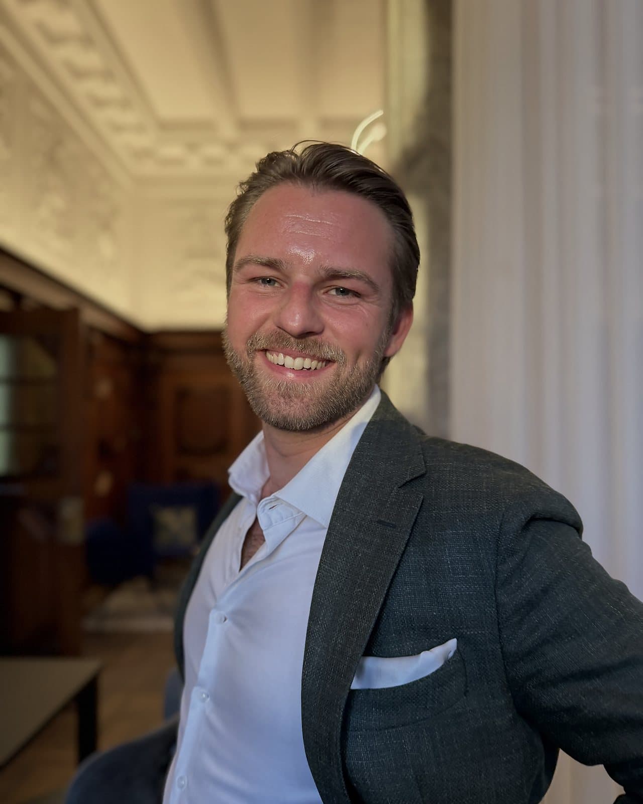 Kim Olsen smiling at the camera in a dark green tweed jacket and white shirt, in a room with an ornate ceiling and white curtains.