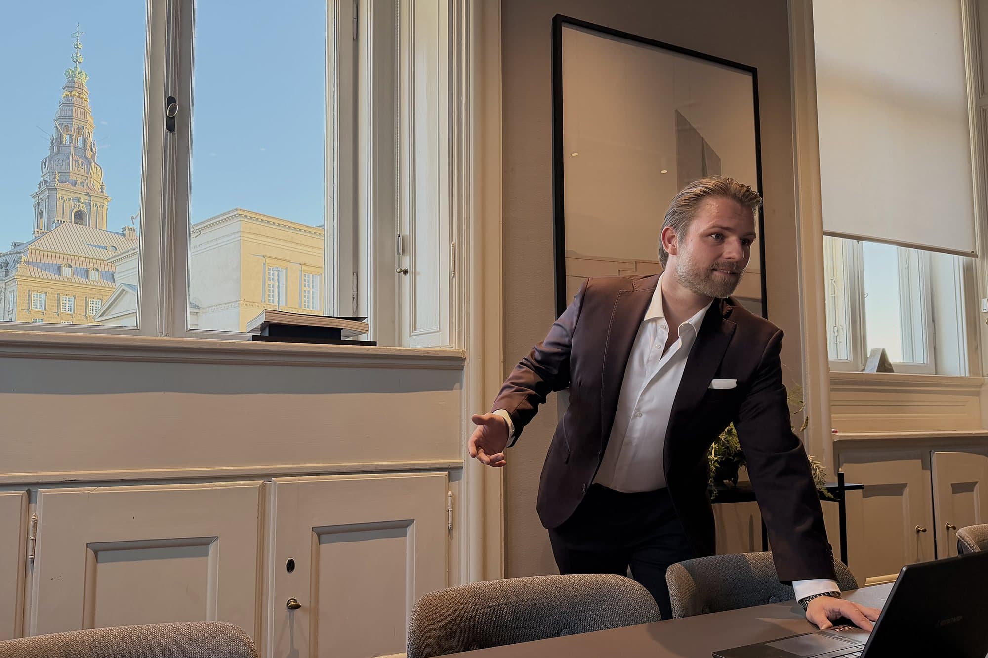 Kim Olsen in a dark suit, leaning on a meeting table and gesturing toward a tall window with an old copper spire outside.